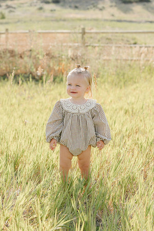 A toddler stands in a grassy field wearing a camel gingham lace bubble romper with a white collar. The soft cotton fabric creates a comfortable look perfect for outdoor play.
