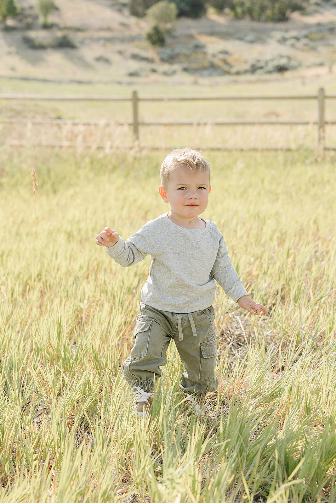 A young child stands in a grassy field, wearing a light gray long-sleeve shirt and green cargo pants.