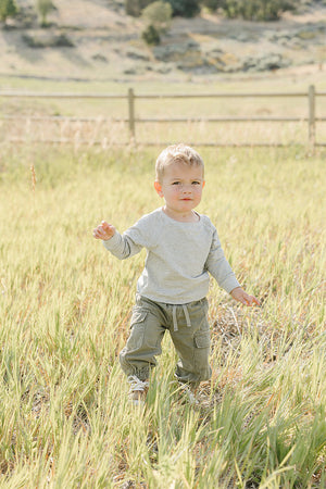 A young child stands in a grassy field, wearing a light gray long-sleeve shirt and green cargo pants.