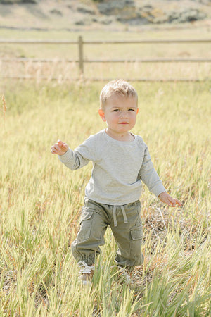 A young child wearing a light grey crewneck sweatshirt and green cargo pants stands in a grassy field, enjoying the outdoors under a clear sky.