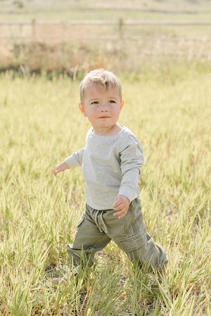 A toddler in a light gray long-sleeve shirt and green cargo pants stands in a grassy field, exploring the outdoors with a curious expression on his face.