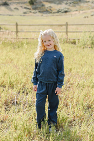 A young girl stands in a grassy field wearing a navy terry cloth tracksuit, featuring a relaxed fit and a small embroidered design. She smiles brightly against a scenic backdrop.