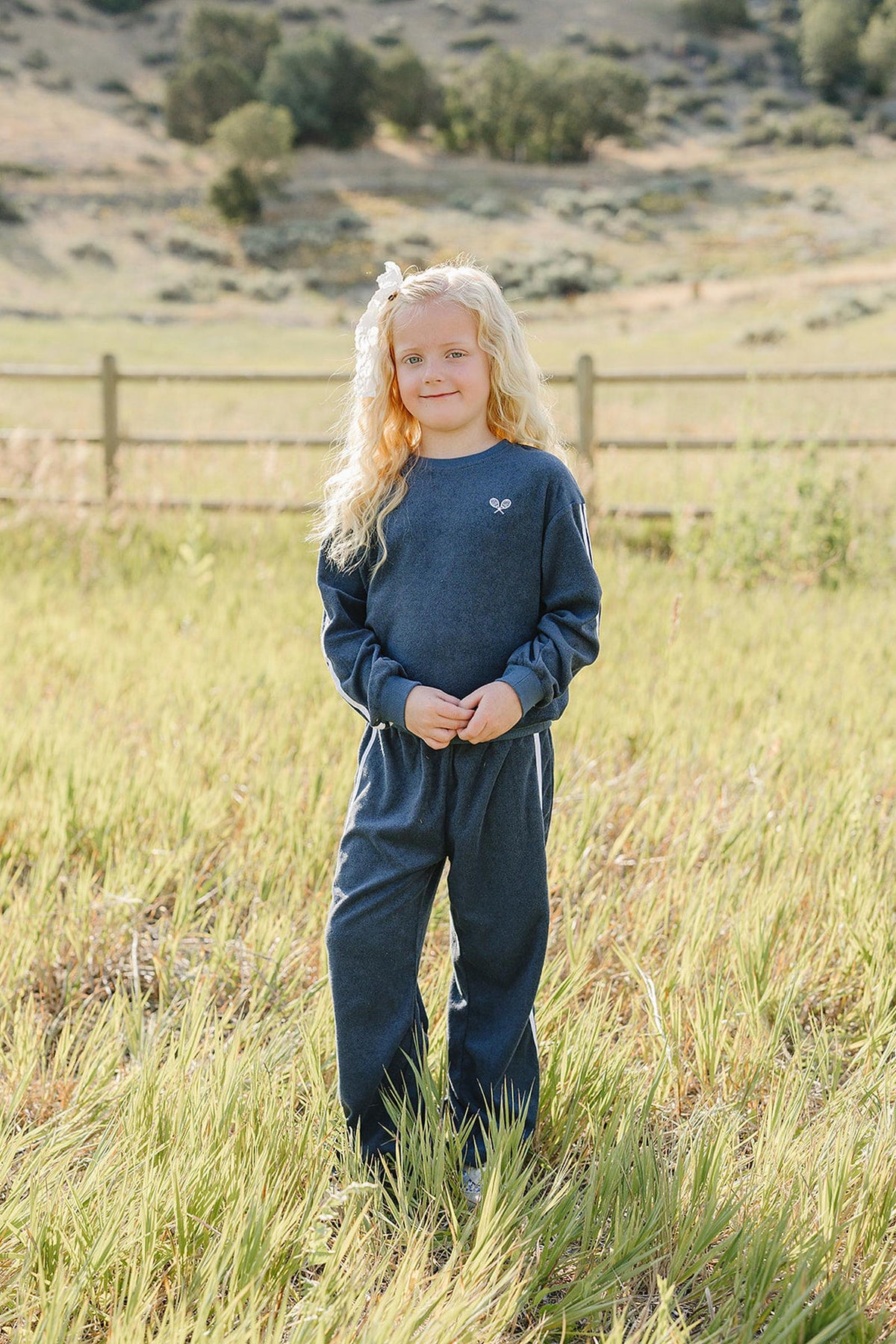 A young girl stands in a grassy field wearing a navy terry cloth tracksuit. The outfit features a relaxed fit and is made from soft, breathable cotton.