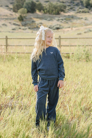 A young girl with blonde hair smiles while wearing a navy terry cloth tracksuit in a grassy field. The outfit features a relaxed fit, perfect for outdoor play and comfort.