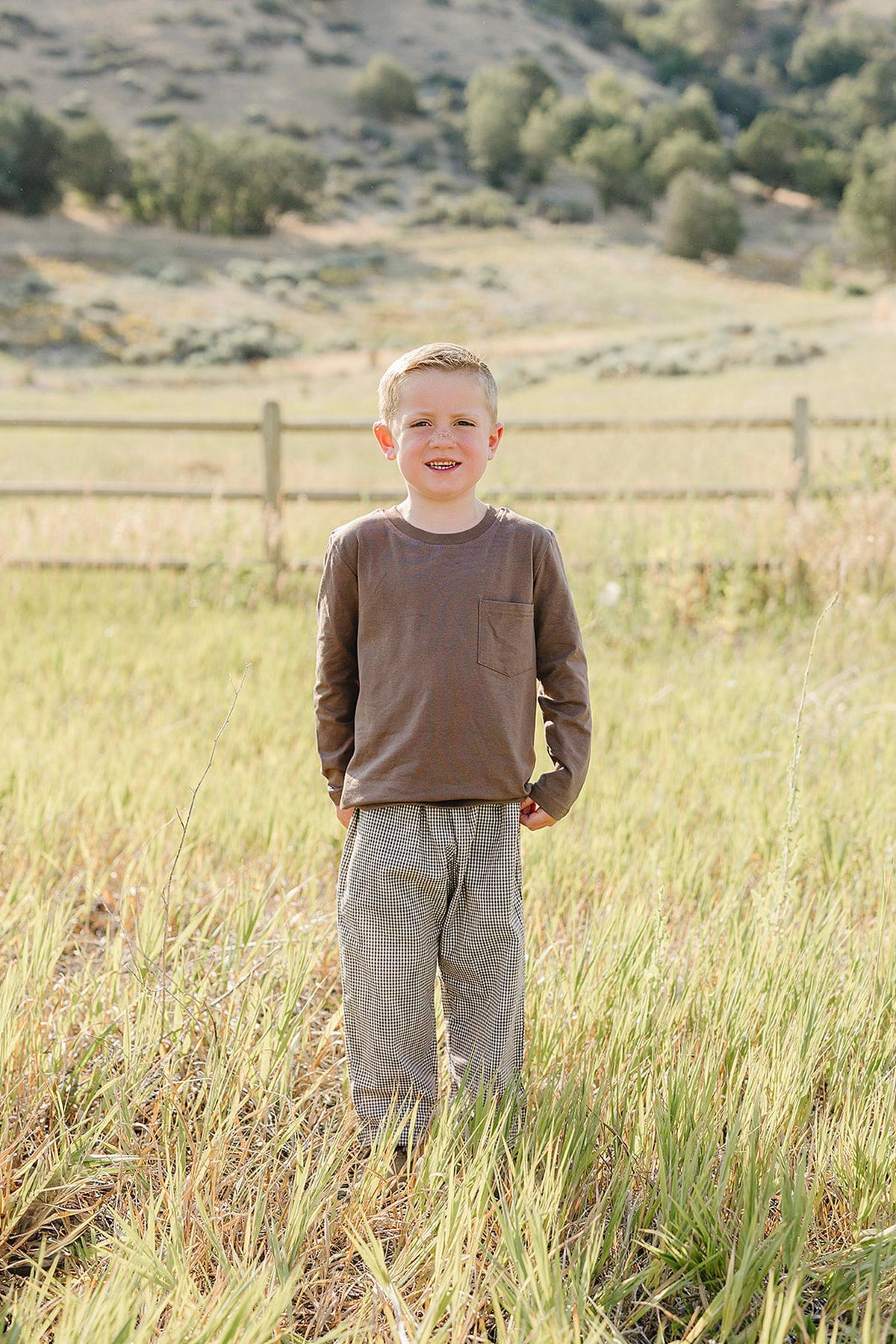 A smiling child stands in a grassy field, wearing a brown long-sleeve shirt and striped cotton pants. The natural setting features rolling hills and a rustic fence in the background.