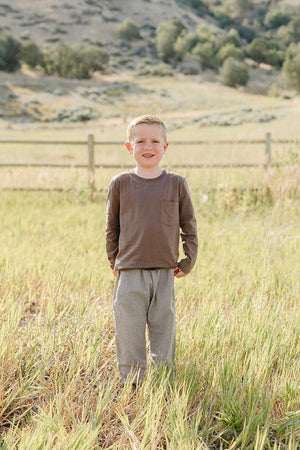 A smiling child stands in a grassy field, wearing a brown long-sleeve shirt and striped cotton pants. The natural setting features rolling hills and a rustic fence in the background.