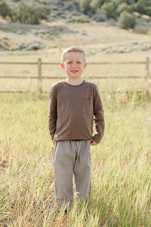 A smiling child stands in a grassy field, wearing a brown long sleeve cotton T-shirt with a pocket and striped pants.