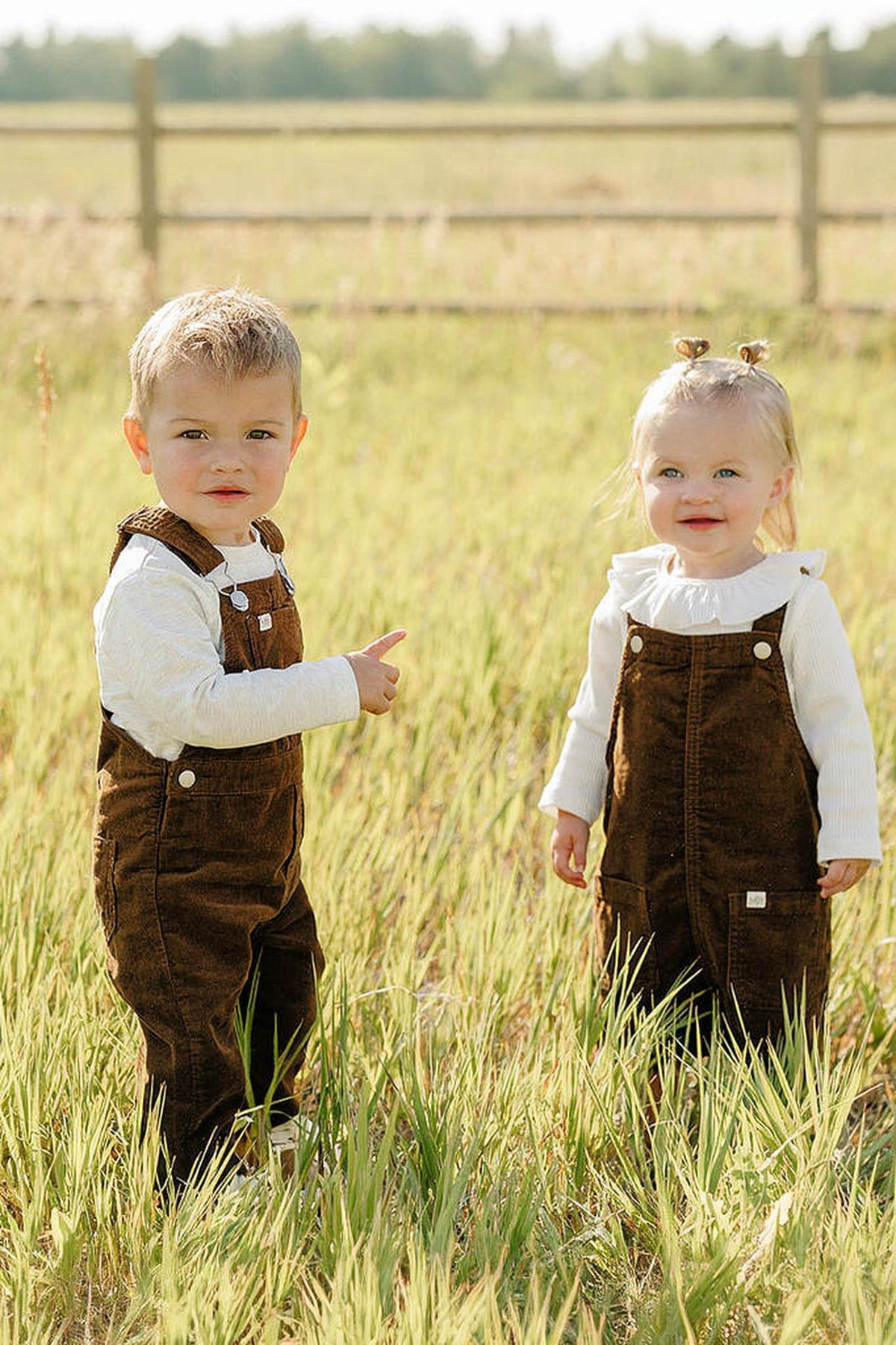 Two young children stand in a grassy field, wearing brown corduroy overalls paired with white long-sleeve shirts. The setting features a wooden fence and a sunny, outdoor backdrop.