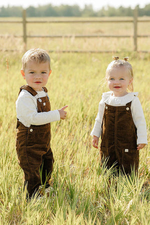 Two young children stand in a grassy field, wearing brown corduroy overalls paired with white long-sleeve shirts. The setting features a wooden fence and a sunny, outdoor backdrop.