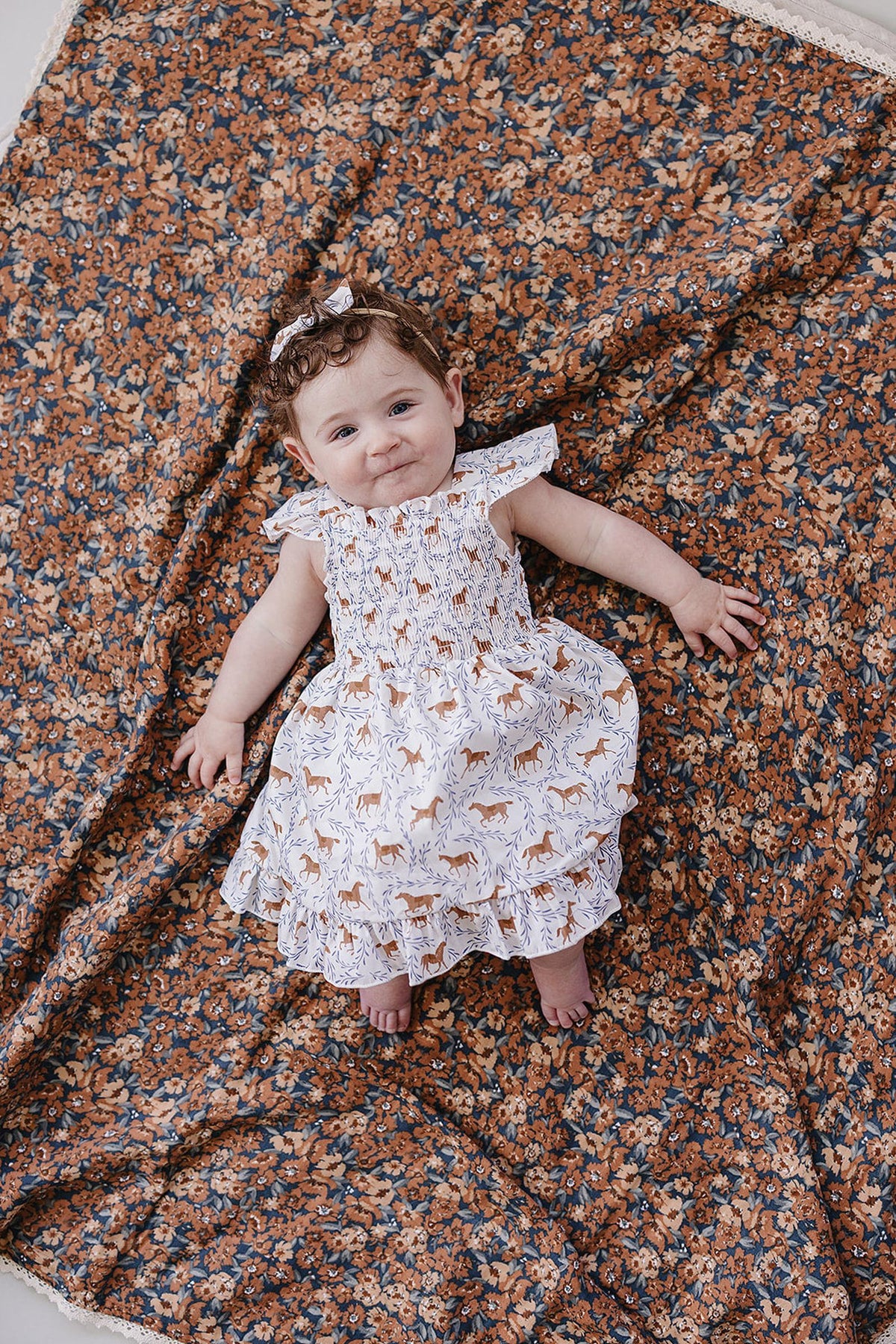 A smiling baby girl lies on a floral-patterned blanket, wearing a white smocked dress featuring playful pony prints.