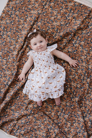 A smiling baby girl lies on a floral-patterned blanket, wearing a white smocked dress featuring playful pony prints.