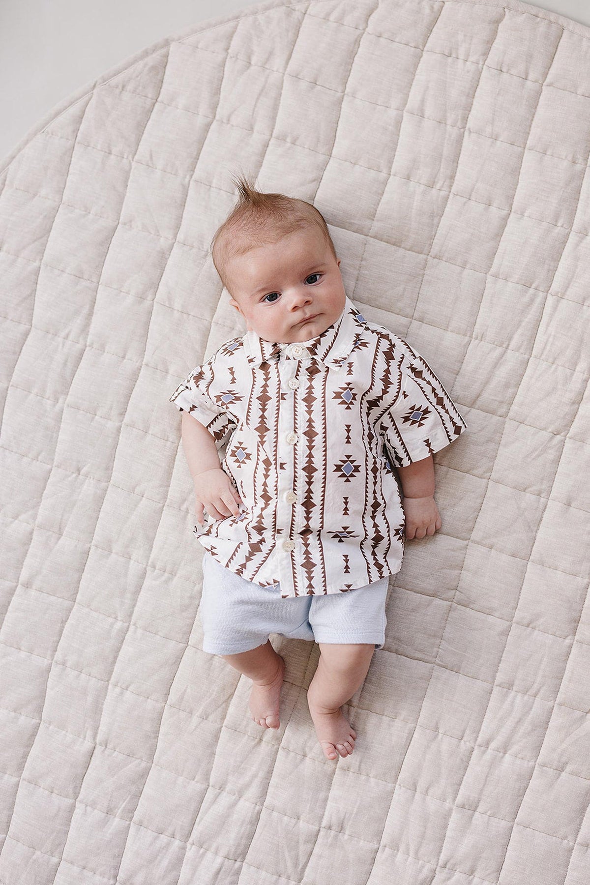 A baby lies on a soft, quilted surface, wearing a white Aztec-patterned button-up shirt with earthy tones and light blue shorts. The shirt features a relaxed fit and playful design.