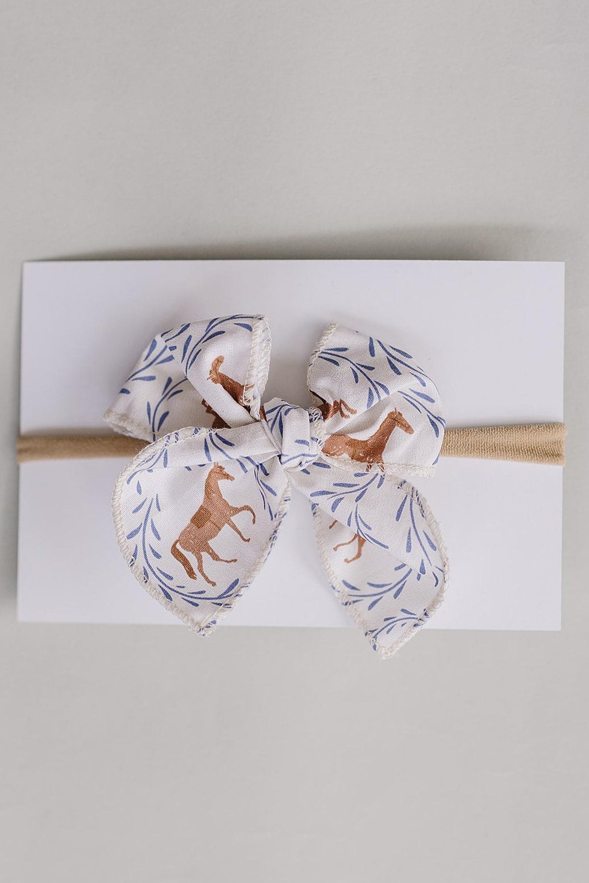 A charming Western pony print hair bow, crafted from soft fabric featuring horses and leaves, is tied neatly on a neutral card with a tan elastic band.