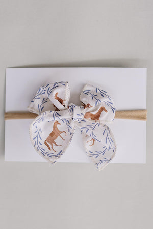 A charming Western pony print hair bow, crafted from soft fabric featuring horses and leaves, is tied neatly on a neutral card with a tan elastic band.