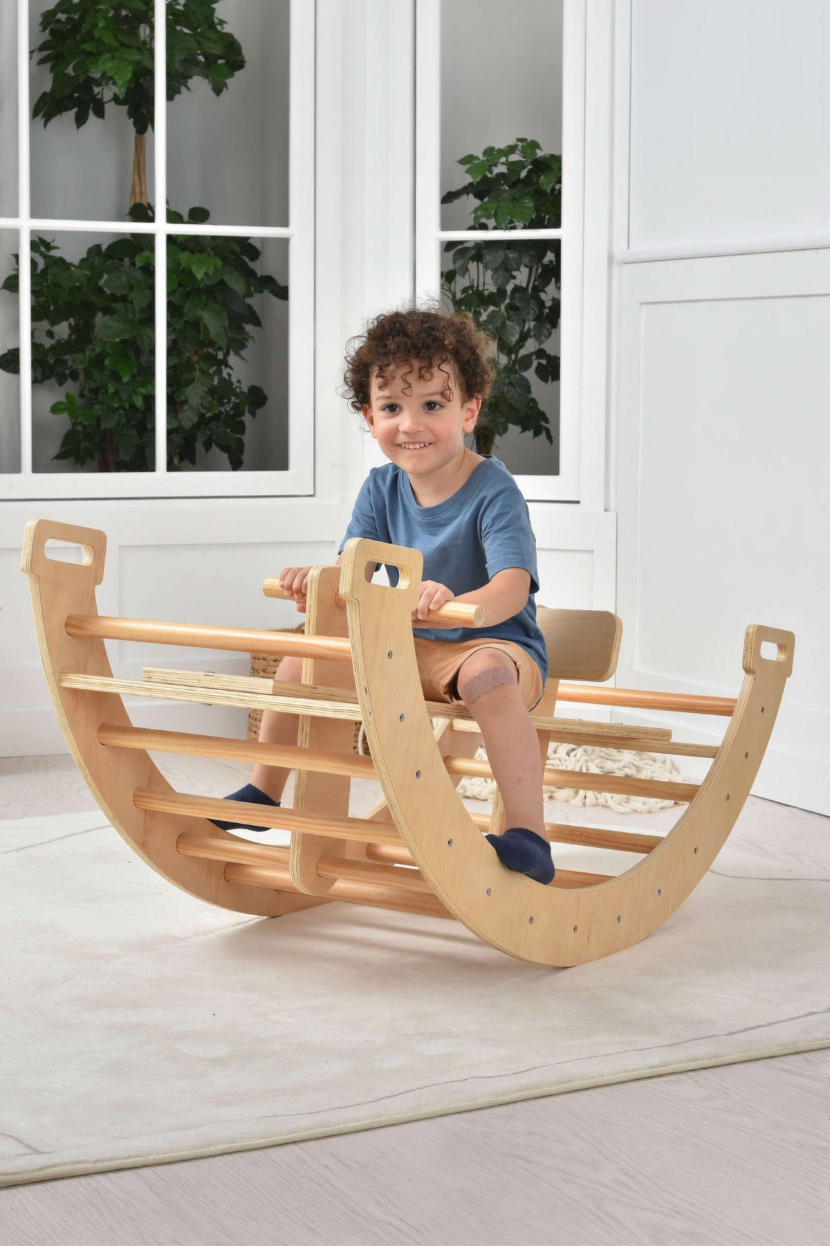 A young boy plays on a wooden Bailis 3 in 1 climbing arch, rocker, and seesaw, smiling while seated in a bright, airy room with plants.