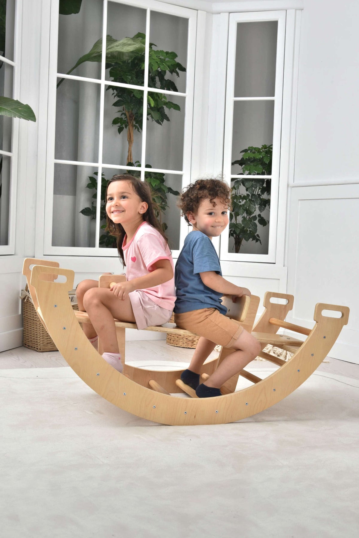Two children are happily playing on a wooden Bailis 3 in 1 climbing arch, rocker, and seesaw, set against a bright, airy room with large windows and greenery.