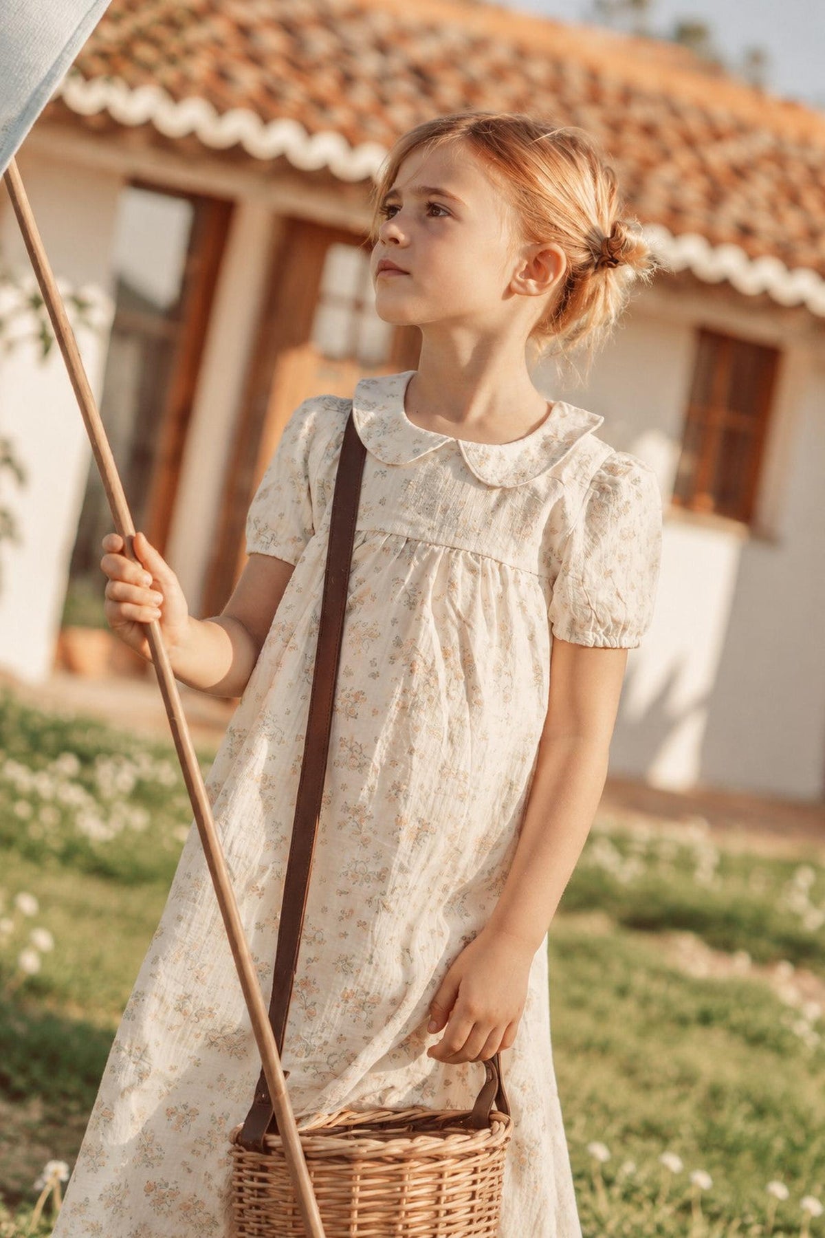 A young girl stands outside in a floral dress with a collar, holding a flag and a woven basket.