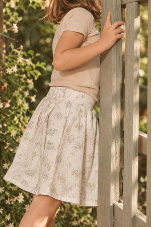 A child wearing a soft, floral-patterned cotton skirt stands beside a wooden structure, surrounded by greenery. The skirt features a light color palette, perfect for casual, stylish outfits.