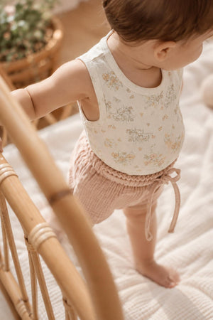 A toddler stands in a cozy nursery, wearing a floral-printed cotton tank top paired with soft, pink bloomers. The setting features natural light and a warm, inviting atmosphere.