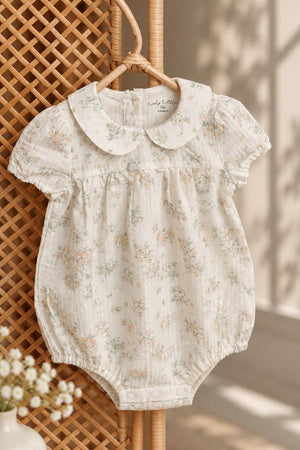 A floral-printed cotton romper hangs against a textured backdrop, featuring a classic Peter Pan collar and delicate ruffle sleeves. Ideal for stylish, comfortable wear for little ones.
