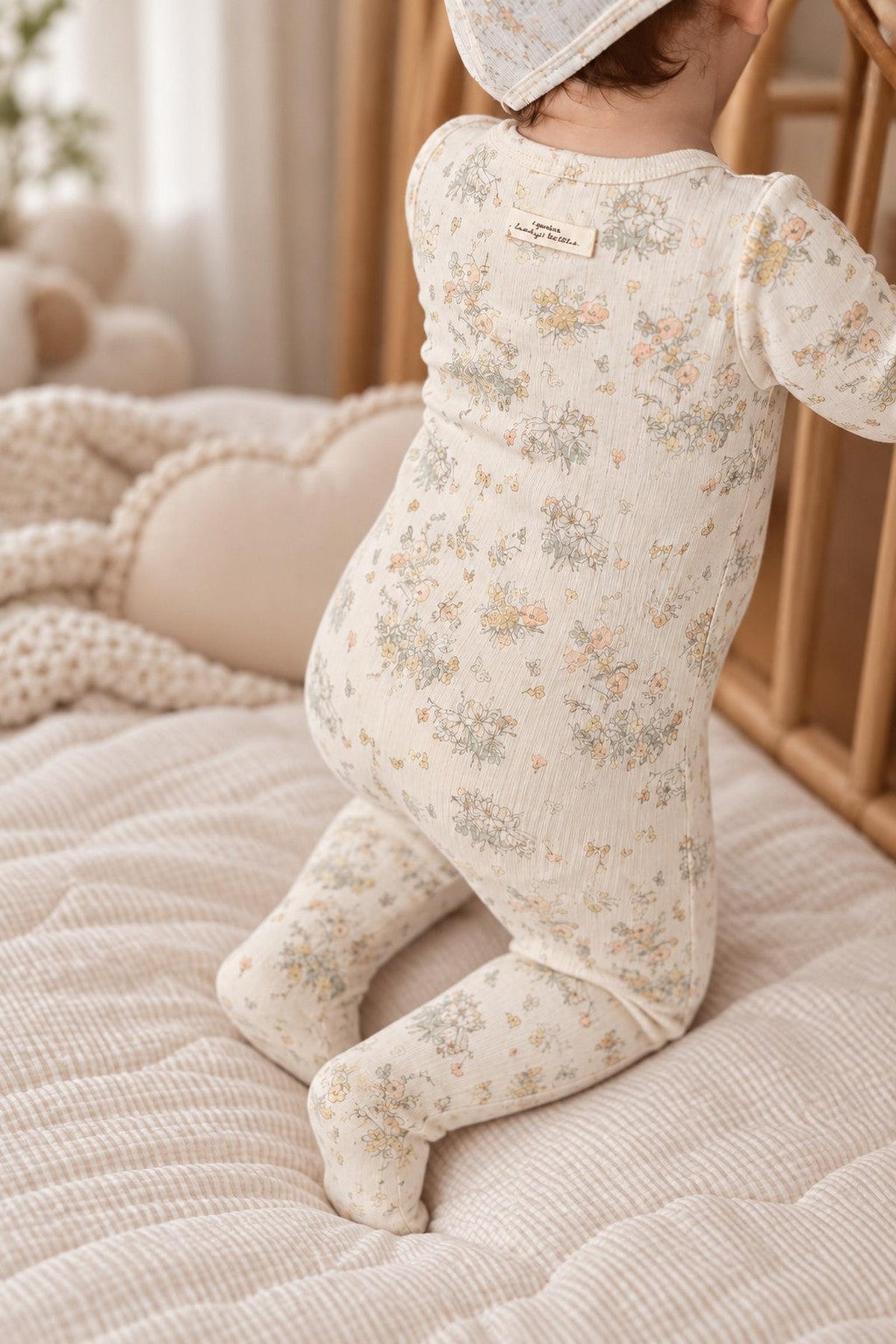 A baby wearing a floral-patterned cotton romper stands on a soft, textured blanket beside a wooden crib. The romper features a gentle color palette and snug fit for comfort.