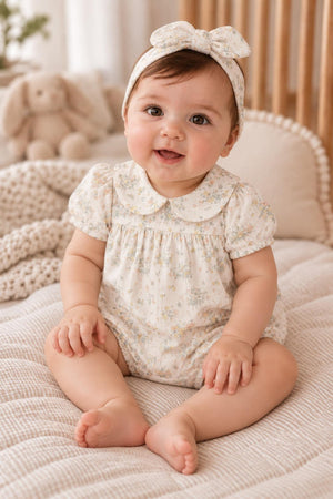 A smiling baby sits on a soft, textured blanket, wearing a floral cotton romper with a Peter Pan collar and a matching headband. The background features a cozy nursery setting.