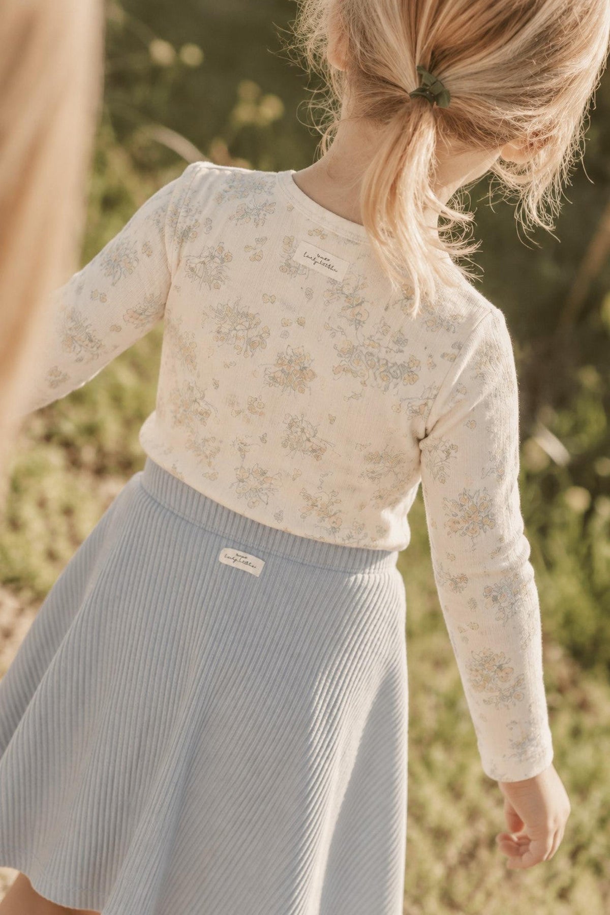 A young girl is seen from behind, wearing a long-sleeve white cotton tee with a floral print and a light blue ribbed skirt, set against a natural outdoor background.
