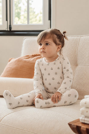 A toddler sits on a light-colored sofa, wearing a cream-colored long-sleeve onesie with small floral prints. The cozy setting features soft cushions and natural light streaming through a window.