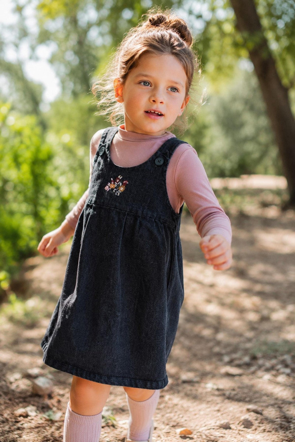 A young girl runs along a sunlit path, wearing a black denim dress with delicate floral embroidery over a pink long-sleeve shirt. The natural setting features greenery.