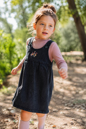 A young girl runs along a sunlit path, wearing a black denim dress with delicate floral embroidery over a pink long-sleeve shirt. The natural setting features greenery.