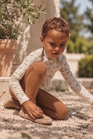 A young child kneels on gravel, wearing a long-sleeved beige top with animal prints and caramel-colored ribbed leggings. The setting features greenery and a sunlit outdoor environment.