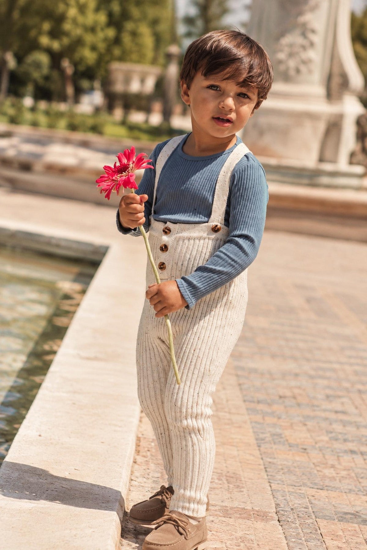 A young boy stands by a fountain, wearing a blue long-sleeve top and cream-colored knit overalls with buttons. He holds a pink flower, showcasing a playful and stylish look.