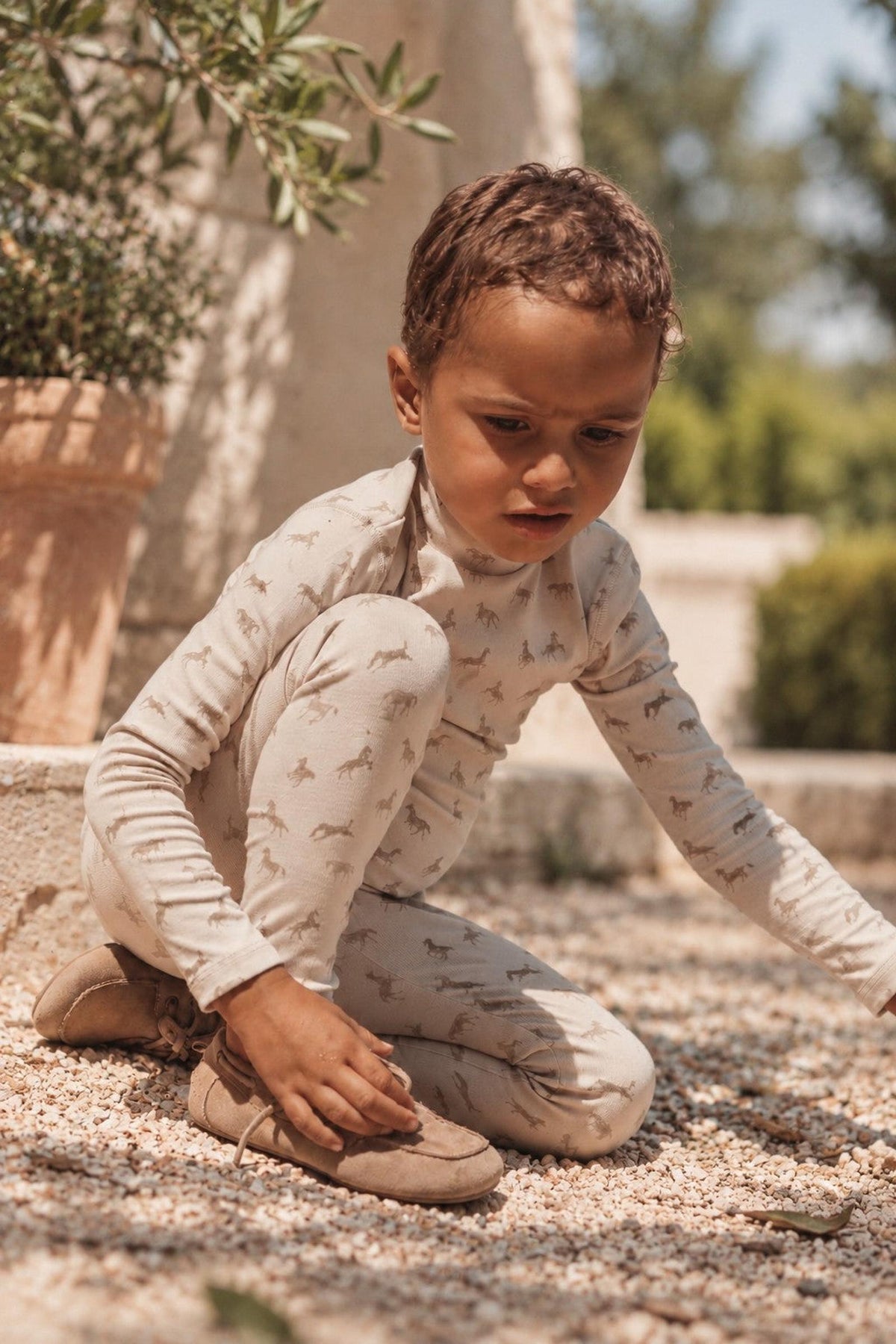 A young child sits on the ground, wearing a light beige outfit with a playful horse print. The setting features natural greenery and soft gravel, highlighting a playful, outdoor atmosphere.
