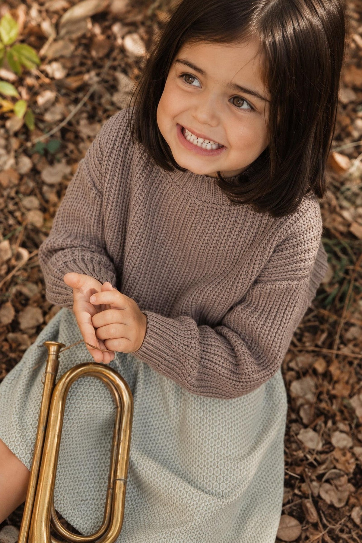 A smiling child sits on the ground surrounded by autumn leaves, wearing a cozy brown sweater and a light green textured skirt, holding a brass trumpet.