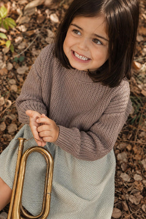 A smiling child sits on the ground surrounded by autumn leaves, wearing a cozy brown sweater and a light green textured skirt, holding a brass trumpet.