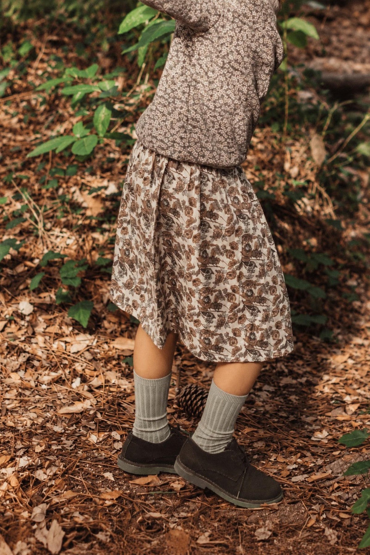 A child wears a brown, floral printed skirt made from soft cotton, paired with cozy socks and shoes, while playing amid lush greenery and fallen leaves in a natural setting.
