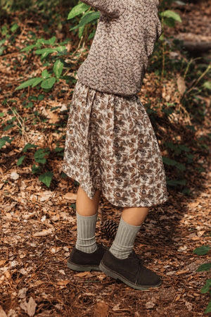 A child wears a brown, floral printed skirt made from soft cotton, paired with cozy socks and shoes, while playing amid lush greenery and fallen leaves in a natural setting.