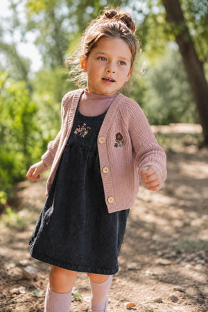 A young girl joyfully walks through a sunny outdoor setting, wearing a mauve chunky knit cardigan with floral embroidery over a dark dress, showcasing a cozy and stylish look for.