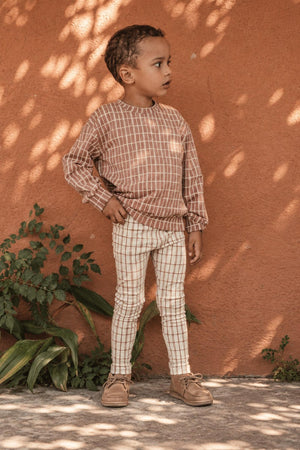 A young child stands against a textured orange wall, wearing a mauve checkered sweater and cream-colored checkered leggings. The outfit is casual and stylish, perfect for everyday wear.