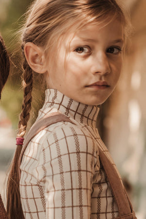 A young girl with braided hair wears a lightweight, check-patterned turtleneck, showcasing a soft fabric and stylish design. The warm, natural lighting enhances the cozy, playful vibe.