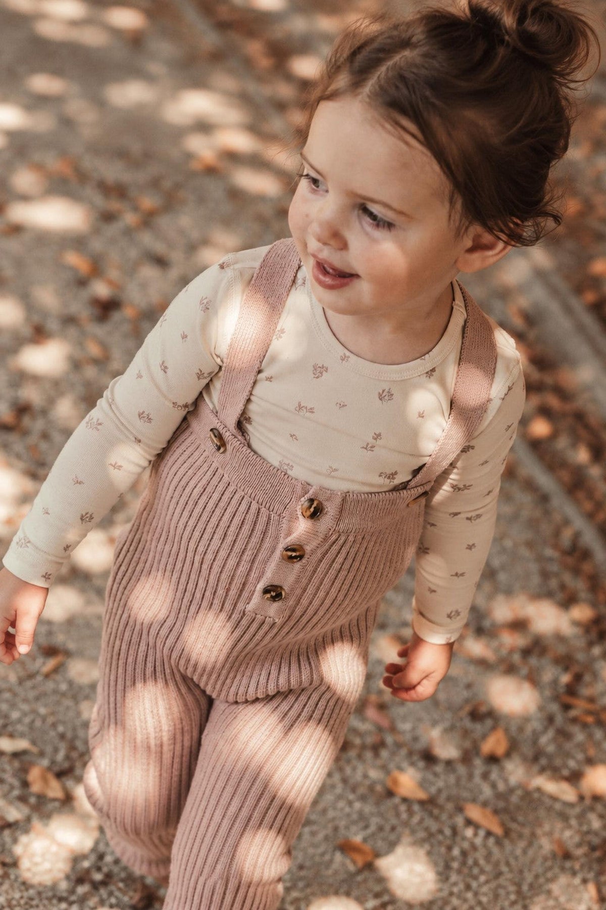 A young child walks outdoors in a mauve knit overall with ribbed texture, featuring button details. She pairs it with a light, patterned long-sleeve shirt, surrounded by fallen leaves.