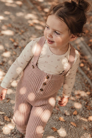 A young child walks outdoors in a mauve knit overall with ribbed texture, featuring button details. She pairs it with a light, patterned long-sleeve shirt, surrounded by fallen leaves.