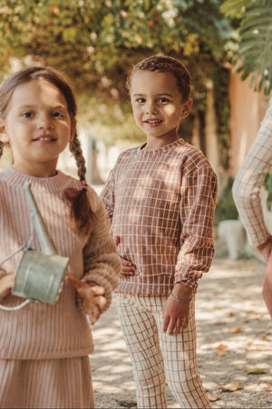 Two children are outdoors, wearing cozy, stylish outfits. One child sports a mauve check printed cotton sweatshirt, while the other wears a matching outfit, surrounded by lush greenery.