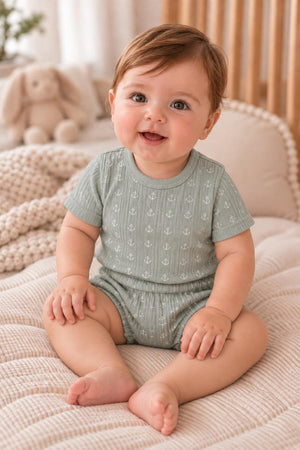 A smiling baby sits on a textured blanket, wearing a light green ribbed onesie with an anchor pattern.