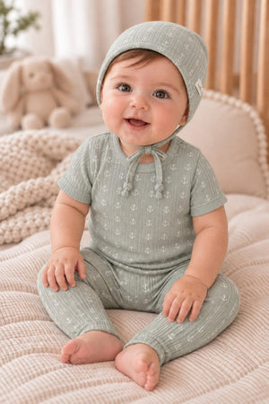 A baby wears a soft, sage green cotton outfit with anchor prints, complemented by a matching bonnet. The cozy nursery setting features plush toys and a textured blanket.