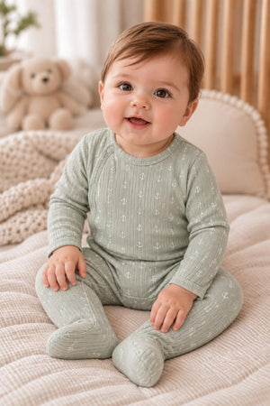 A smiling baby sits on a soft, neutral-toned blanket, wearing a light green ribbed romper with anchor prints. The cozy setting includes a plush teddy bear in the background.
