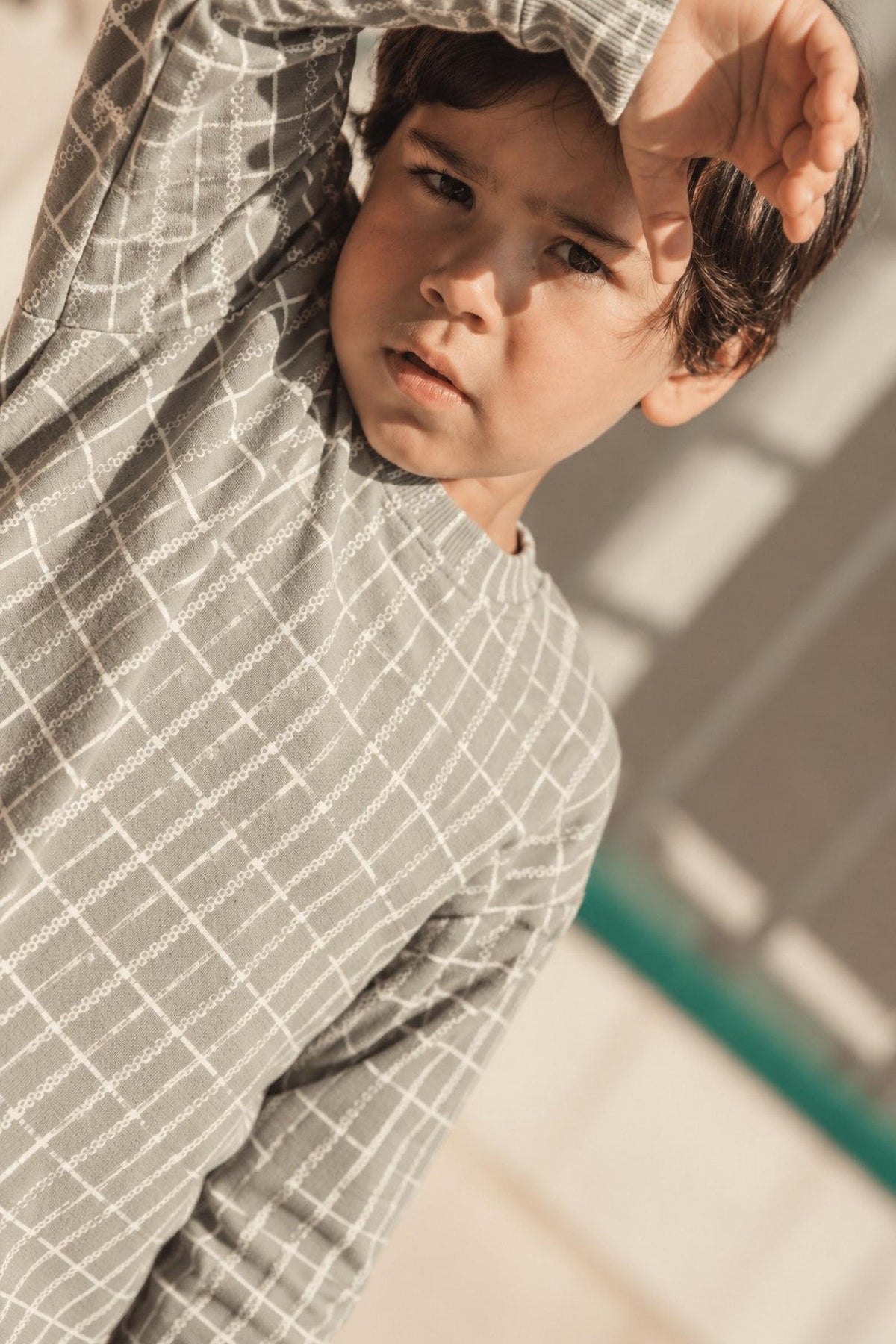 A young boy wearing a sage green check printed sweatshirt stands in natural light, looking thoughtfully at the camera.
