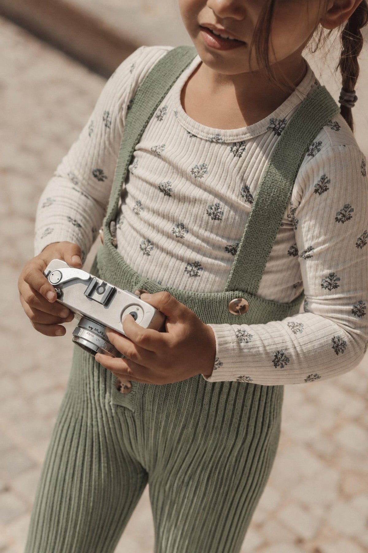 A young child holds a vintage camera while wearing sage green knit overalls and a long-sleeve striped shirt with floral patterns. The outfit combines comfort and style for playful moments.