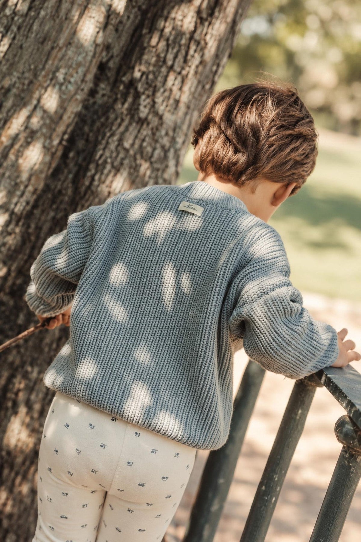 A child in a cozy sage chunky knit cotton sweater leans against a wooden railing, surrounded by trees and greenery. The sweater features a relaxed fit, perfect for outdoor play.