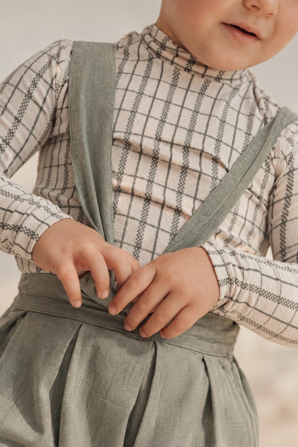 A child is wearing a sage check printed long sleeve cotton turtleneck paired with a stylish, light-colored suspender outfit, showcasing a cozy and playful look perfect for kids.
